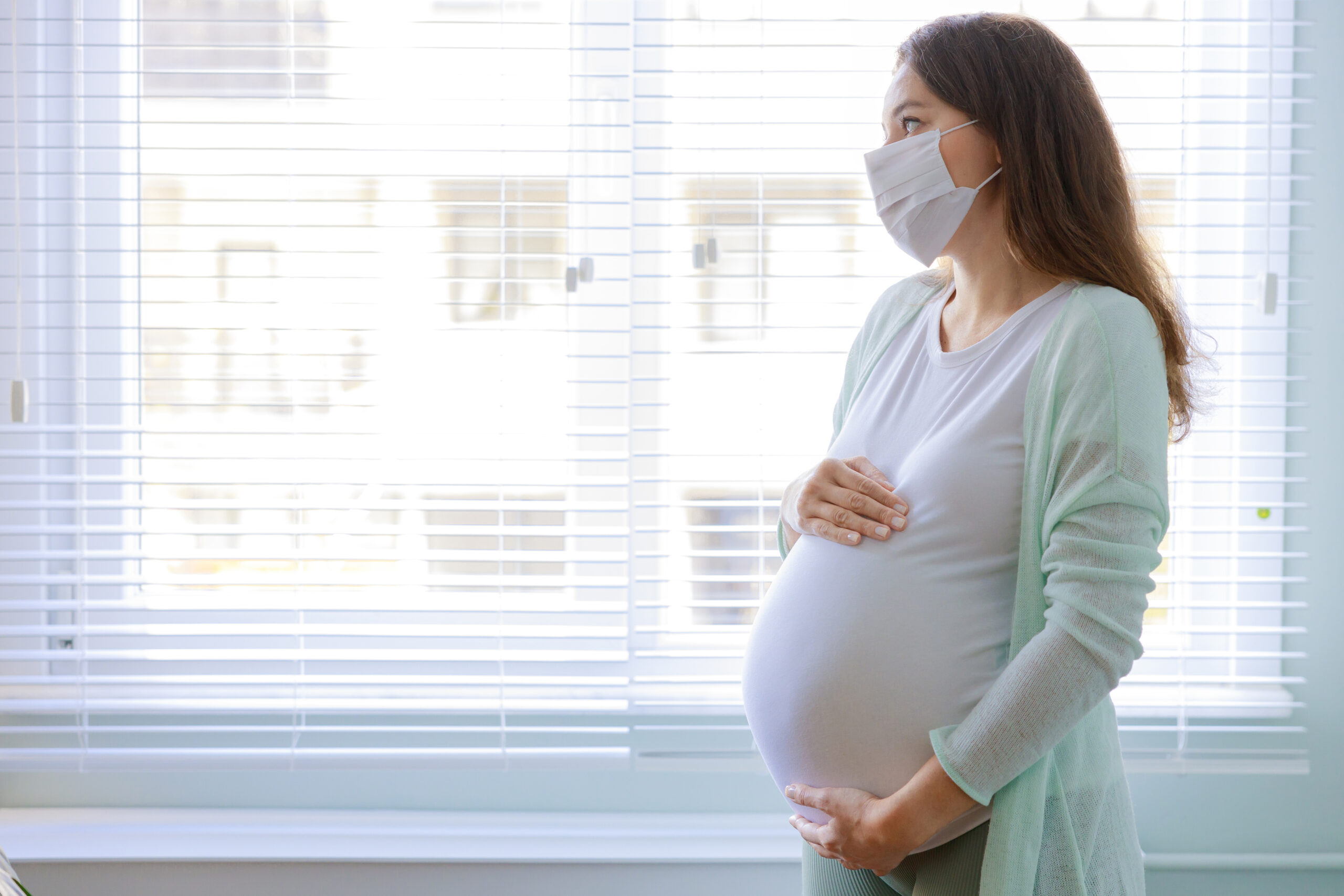 Pregnant person wearing surgical mask stands by window.