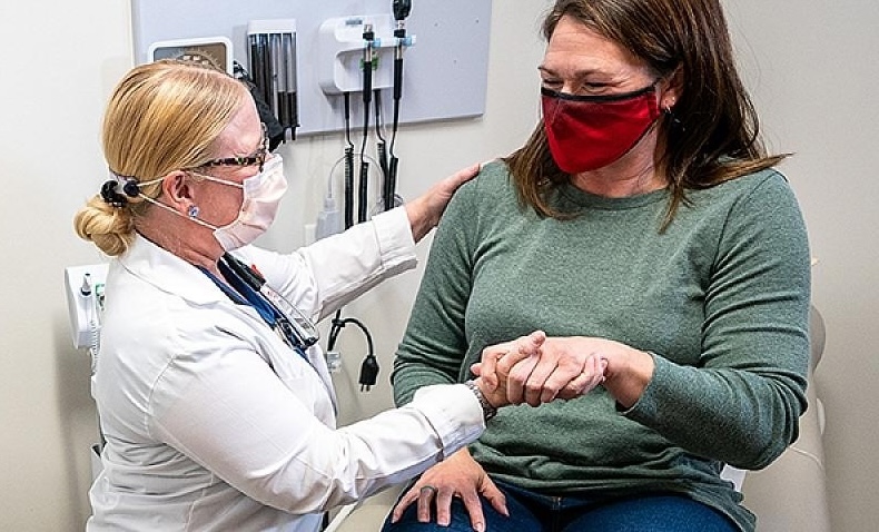 A provider in a white coat helps a seated patient. Both are masked.